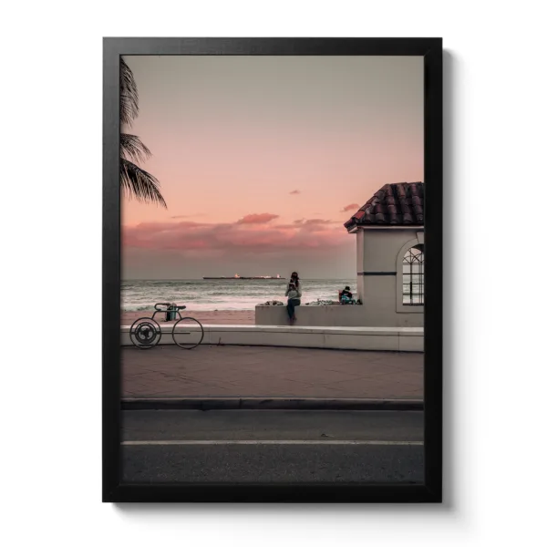 Photograph of a sunset where a woman is sitting on the kerb reading a book. In the background is the sea and a small white hut.