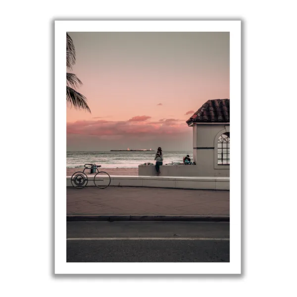 Photograph of a sunset where a woman is sitting on the kerb reading a book. In the background is the sea and a small white hut.