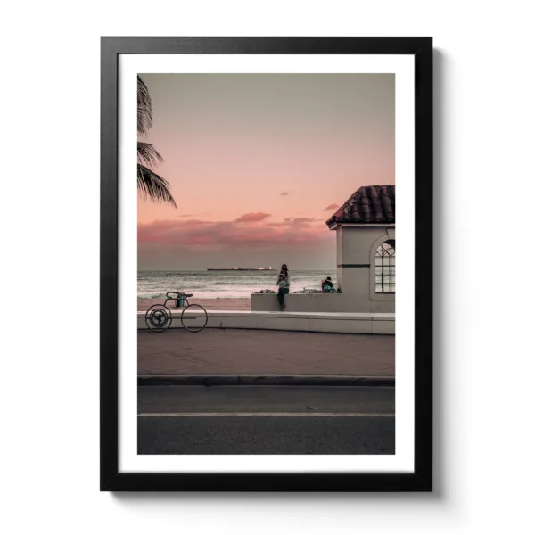 Photograph of a sunset where a woman is sitting on the kerb reading a book. In the background is the sea and a small white hut.