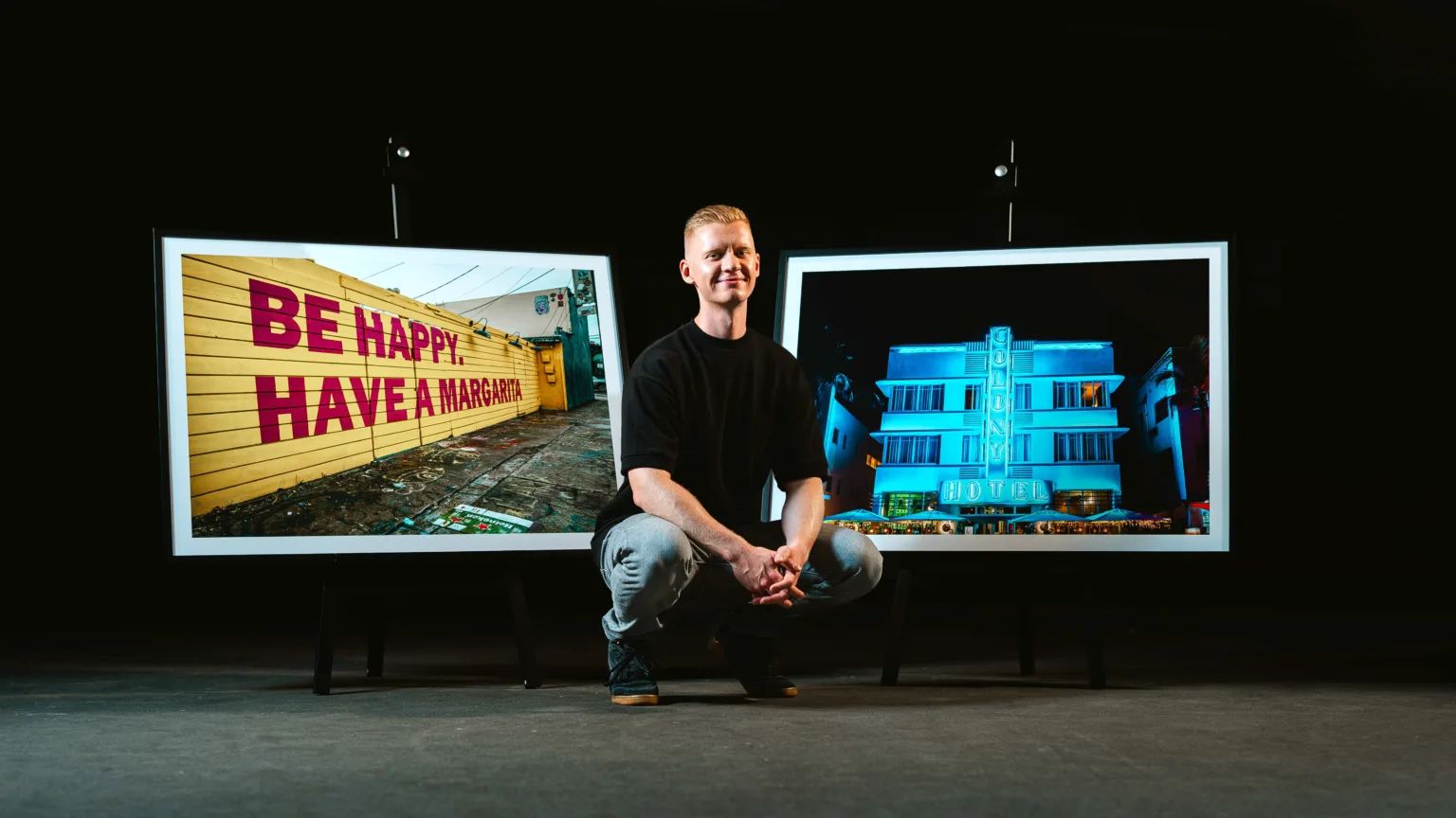 Mann hockt lächelnd zwischen zwei gerahmten Fotos: links gelbe Wand mit „BE HAPPY. HAVE A MARGARITA“, rechts das beleuchtete „Colony Hotel“ bei Nacht.