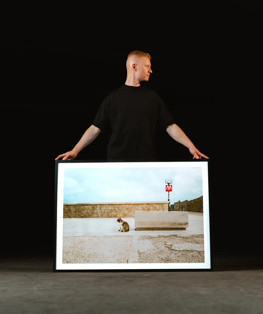 Mann in schwarzem T-Shirt hält einen großen gerahmten Fotodruck mit einer Katze vor einer Steinmauer und einem Metro-Schild.